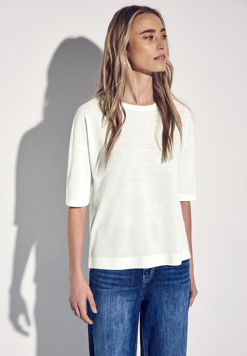 Hvid, løs pasform t-shirt med korte ærmer, lavet af et let materiale; har en rund halsudskæring og let tekstur. Bæres med blå jeans.