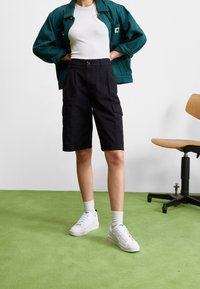 Veste verte, débardeur blanc, shorts cargo noirs, baskets blanches et chaussettes mi-mollet. Tissu texturé avec poches cargo et fermeture à bouton.
