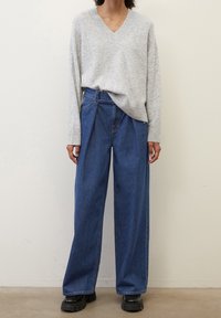 Maglione grigio chiaro a V con polsini a coste, abbinato a jeans blu a vita alta e gamba larga. Il modello indossa stivali neri con plateau spessi.