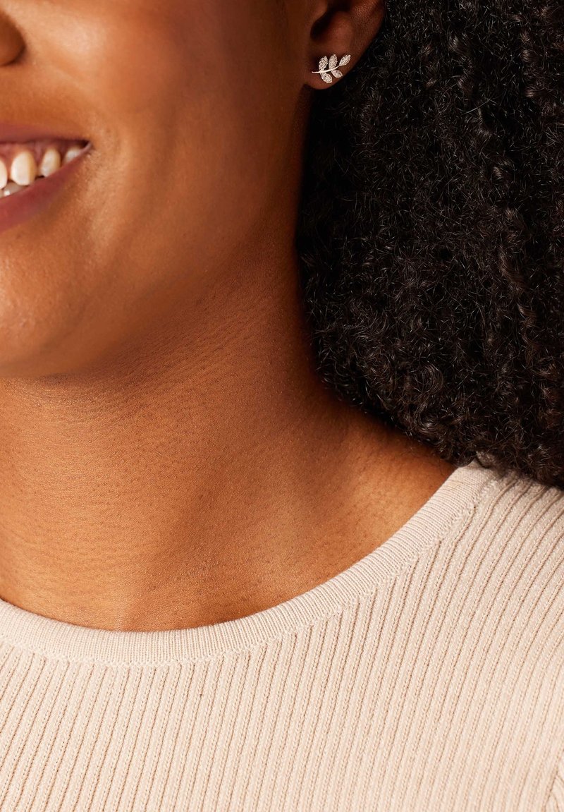 Silberne, blattförmige Ohrringe an einem Modell, die über eine filigrane Detailgestaltung und eine strukturierte Oberfläche verfügen. Das Modell trägt ein geripptes, helles Oberteil.