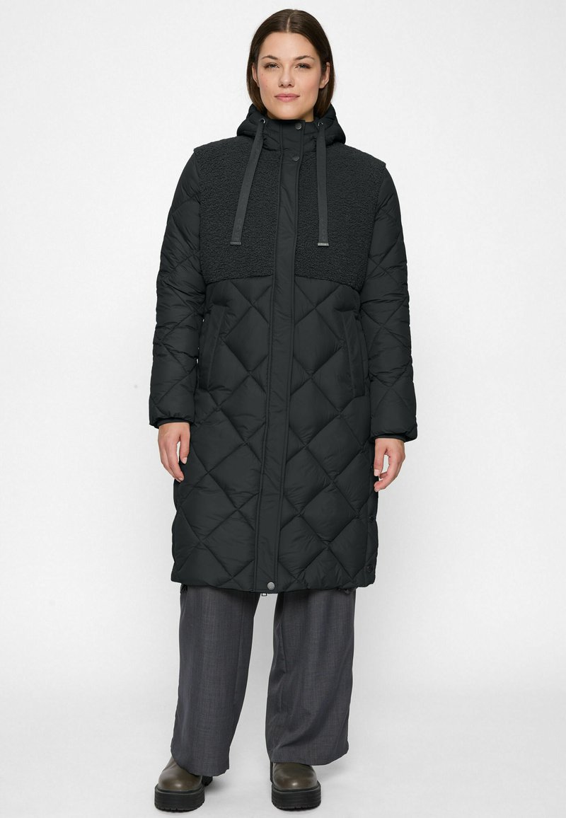 Woman wearing a long black quilted winter coat with a hood and textured upper panel, gray wide-leg pants, and dark boots against a white background.