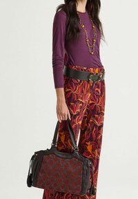 Borsa nera con motivo rosso e nero texturizzato, manici robusti e accessori decorativi. Modella con top viola e pantaloni fantasia.