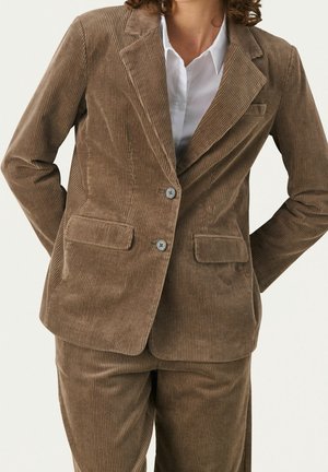 Frau trägt einen braunen Cord-Anzug mit einem Zweiknopf-Blazer über einem weißen Hemd mit Kragen, steht mit den Händen hinter dem Rücken.