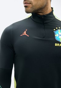 Homme portant un haut de sport noir avec des accents jaunes, un écusson de l'équipe du Brésil et un logo Jumpman rouge sur la poitrine.