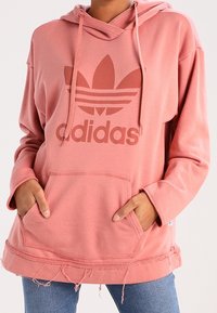 Růžová mikina Adidas s velkým logem, klokaní kapsou, kapucí na šňůrku a detailním zakončením s třásněmi. Vyrobena z měkkého a pohodlného materiálu.
