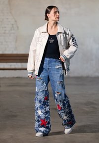 Bijela bomber jakna s vezom zvjezdica, crni majica bez rukava s logom, i izlizane plave traperice s crvenim i bijelim zvjezdastim zakrpama. Bijele tenisice.