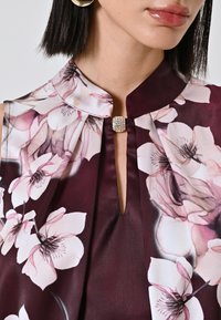 Blusa in satin color bordeaux con colletto alto, caratterizzata da una stampa floreale e un dettaglio a occhio di chiave sullo scollo, accentuata da una chiusura con gioiello.