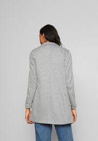 Mujer con cabello largo y oscuro viste una chaqueta gris a rayas y jeans azules, de pie frente a un fondo gris claro liso, con la espalda hacia la cámara.