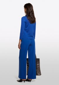 Blå stickad långärmad topp och matchande vida byxor. Svart axelväska i handen. Slät textur, avslappnad design och enkel silhuett.