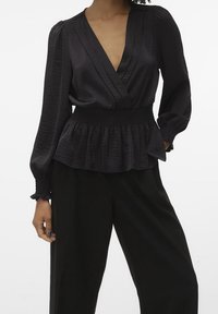 Blouse en peplum noire avec un décolleté en V profond, des manches longues, une taille élastique et un tissu texturé, associée à un pantalon large noir.