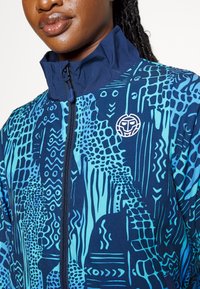 Marine blauwe zip-up jack met levendige abstracte patronen, hoge kraag en een logo patch. Gemaakt van gladde, rekbare stof.
