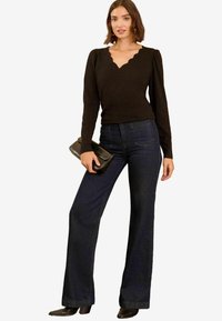 Fekete, hosszú ujjú felső, hullámos nyakkivágással, sötét farmerekkel kombinálva, amelyen a szárak szélesednek. A modell egy fekete clutch-ot tart a kezében. Sima anyagú, testhezálló fazon.
