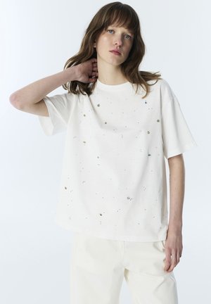 Femme aux cheveux bruns mi-longs portant un t-shirt blanc oversize avec de petites taches multicolores, posant avec une main derrière le cou.