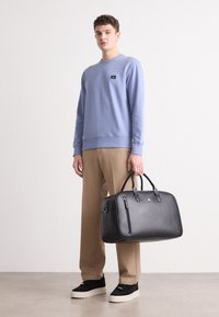 Sort læder weekendtaske med to håndtag, der har en yderlomme med lynlås og en glat tekstur. Modellen bærer en lyseblå sweatshirt og beige bukser.