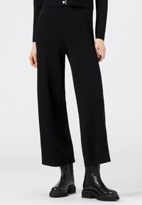 Černé široké kalhoty vyrobené z hladké látky, s vysokým pasem a minimalistickým designem. V kombinaci s masivními černými kotníkovými botami.