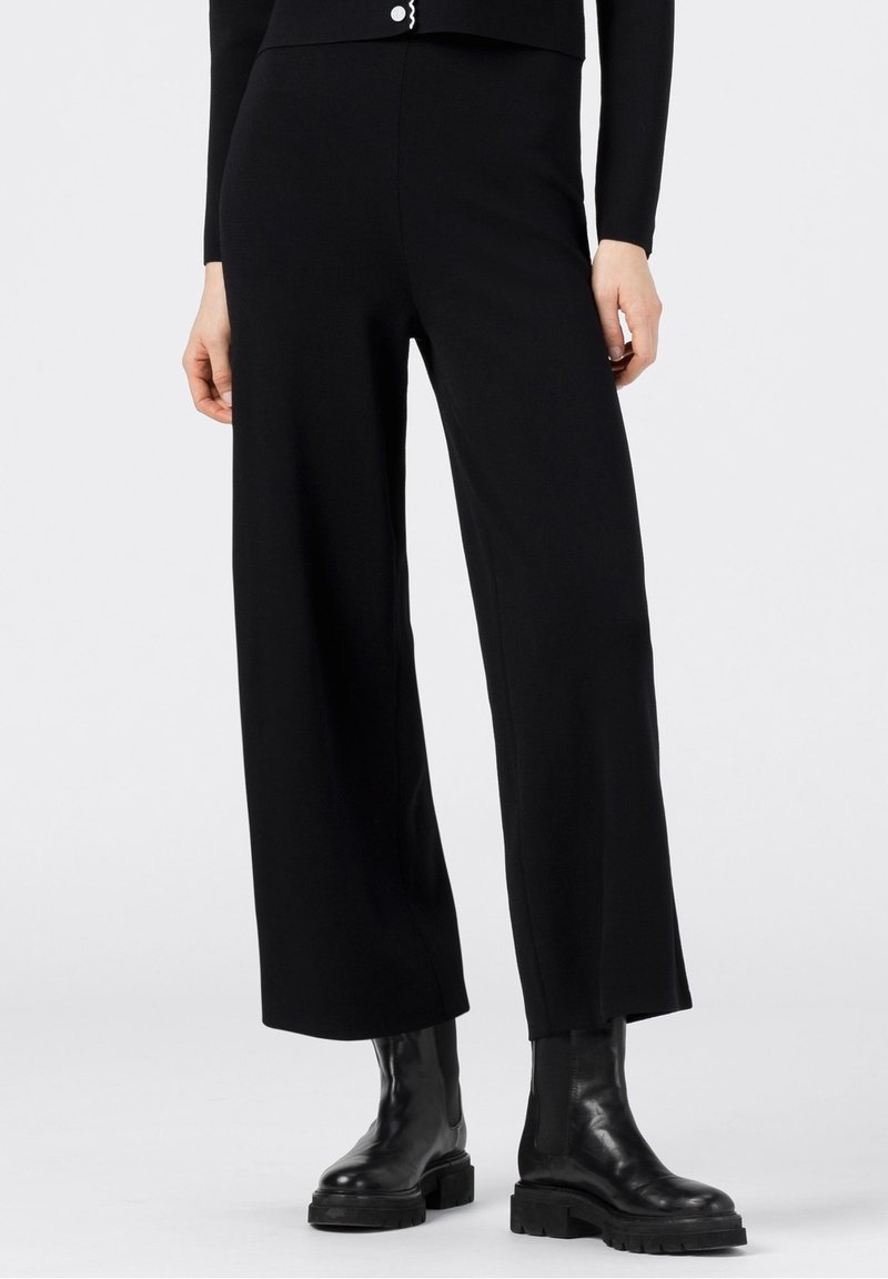 Černé široké kalhoty vyrobené z hladké látky, s vysokým pasem a minimalistickým designem. V kombinaci s masivními černými kotníkovými botami.