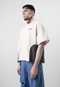Hellbeige oversized T-Shirt mit einem schwarzen, strukturierten Einsatz. Verfügt über ein kleines Logo auf der Brust und rohe Kanten für ein einzigartiges Design.
