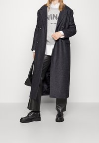 Abrigo de lana gris oscuro largo con frente de botones dobles, pantalones de cuero negros, sudadera gris con texto "MILANO" y zapatos de cordones negros.