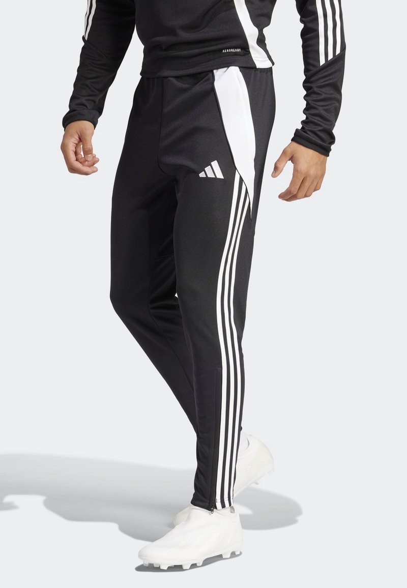 Homme portant un pantalon de survêtement Adidas noir avec des bandes blanches le long des jambes et des chaussures de sport blanches, debout devant un fond clair.