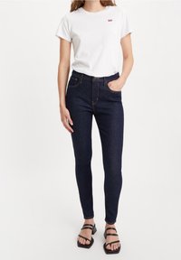 Camiseta blanca de manga corta con un pequeño logo rojo, combinada con jeans ajustados azul oscuro y sandalias negras de tira. Diseño y ajuste simple.