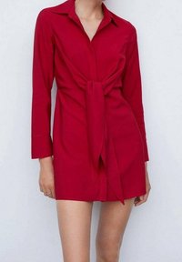 Robe chemise rouge avec col, manches longues et détail noué à l'avant. Tissu lisse et silhouette ajustée, arrivant à mi-cuisse.