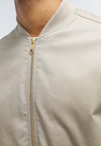 Gros plan sur une veste beige avec une fermeture éclair dorée et un col côtelé portée par une personne.