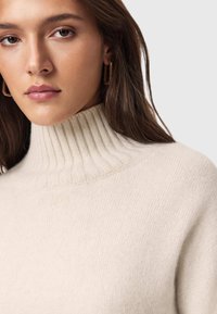 Kremowy sweter dzianinowy z wysokim prążkowanym dekoltem, miękką fakturą i subtelnymi kropeczkami. Modelka nosi złote geometryczne kolczyki w formie kół.