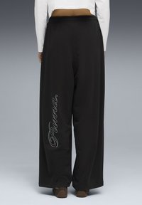 Άτομο που φοράει μαύρο παντελόνι με φαρδιές παντελόνες με την επιγραφή "Proenza" γραμμένη κάθετα στο αριστερό πόδι, λευκό μακρυμάνικο τοπ και καφέ παπούτσια, στέκεται γυρισμένο προς τα πίσω.