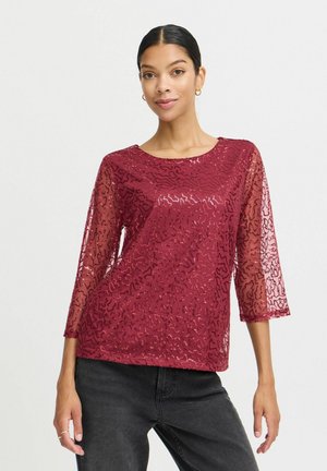 Burgundyröd genomskinlig topp med abstrakt mönster, rund halsringning och trekvarts ärmar, kombinerad med svarta jeans. Texturerad, lättviktsman. 