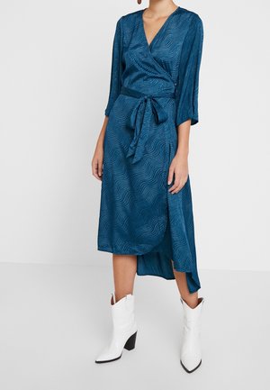 Femme portant une robe portefeuille bleu sarcelle avec des motifs subtils en forme de vague et des bottes blanches à talons mi-mollet, debout sur un sol gris clair.