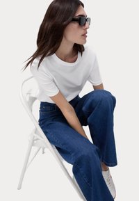 Hvid kortærmet t-shirt, løse blå denim jeans og hvide sneakers. Sorte rektangulære solbriller. Sidder på en hvid stol.