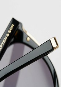 Musta aurinkolasit kiiltävällä pinnalla, joissa on kultaiset yksityiskohdat sangan kohdalla ja kaiverrettu brändin nimi. Linssit ovat tummat ja hieman sävytetyt.