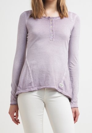 Kobieta ubrana w wyblakłą lawendową długorękawową koszulę henley z guzikami i białe spodnie na jednolitym tle.