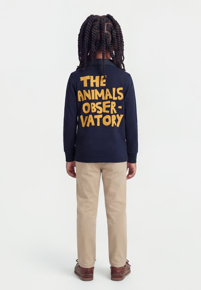 Enfant aux cheveux longs et tressés, debout de dos, portant une chemise bleu marine avec l'inscription "THE ANIMALS OBSERVATORY" en grosses lettres jaunes.