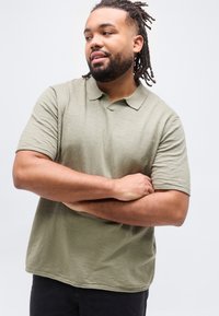 Άνδρας με γενειάδα και ντίντλοκς, φορώντας ανοιχτό πράσινο πουκάμισο πόλο, κοιτάει προς τα πλάγια με σταυρωμένα χέρια μπροστά σε ένα απλό λευκό φόντο.