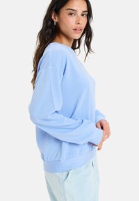 Sweatshirt bleu clair à manches longues, coupe décontractée, encolure large, ourlet élastique et texture lisse. Porté sur un pantalon de couleur claire.