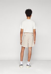Calções bege feitos de uma mistura de algodão, com bolsos traseiros. Combinados com uma t-shirt branca e ténis cinza com riscas. Vista de trás.