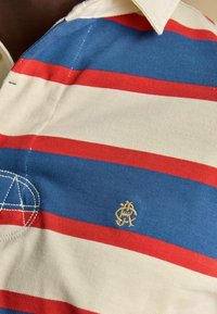 Raidoja polo-paita sinisessä, punaisessa ja kerma-nuanssissa, jossa on pieni kultalangasta brodeerattu emblem ja vaalea kaulus tummalla iholla.