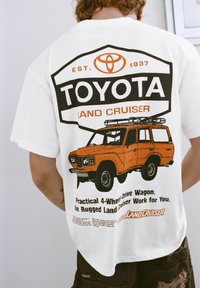 Biała koszulka z dużą czarno-pomarańczową grafiką Toyota Land Cruiser, zawierającą tekst o jej praktyczności i wytrzymałej konstrukcji.