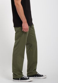 Olijfgroene broek van stevig materiaal, met een rechtvallend ontwerp en subtiel logodetail vlak bij de taille, gecombineerd met zwarte sneakers.