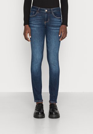Person iført mørkeblå slim fit jeans, sort langærmet top og sorte chunky loafers, stående mod en ensartet lys baggrund.