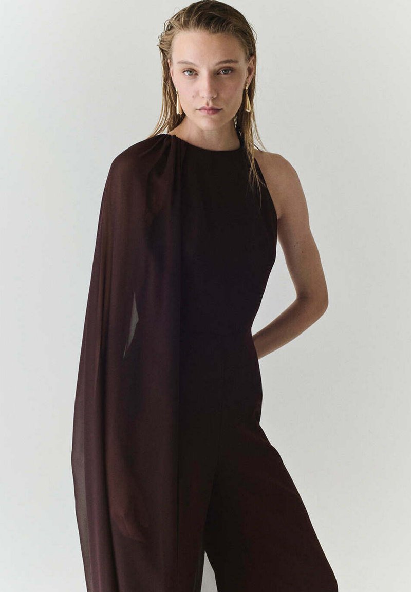 Femme aux cheveux mouillés portant une combinaison noire sans manches avec une cape transparente sur un côté et de longues boucles d'oreilles dorées, debout devant un fond uni.
