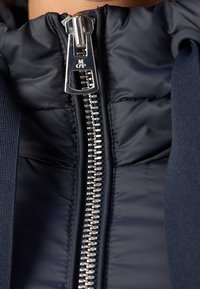 Chaqueta de plumas azul marino con cremallera plateada central, textura acolchada y tela contrastante en los laterales. Cuello forrado con capucha adicional.