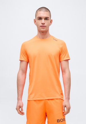 Junger Mann mit kurzen Haaren trägt ein hellorangefarbenes Sportshirt der Marke Borg und passende Shorts, steht vor einem schlichten hellen Hintergrund.