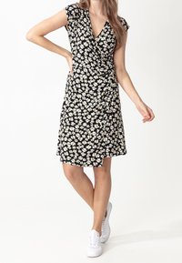 Kobieta w czarnej, sięgającej kolan sukience kopertowej w białe kwiaty, ubrana w białe trampki, pozuje z ręką na biodrze na białym tle.