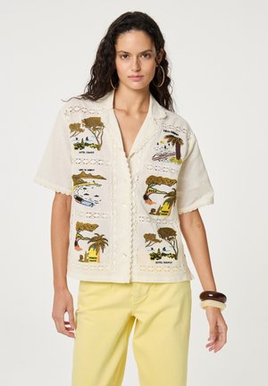 Vrouw met lang krullend haar, gekleed in een crèmekleurig overhemd met tropische scenes geborduurd en gele broek, staand tegen een witte achtergrond.