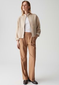 Giubbotto bomber beige in misto lana con chiusura a zip e due tasche frontali, abbinato a pantaloni su misura marrone chiaro e scarpe nere.