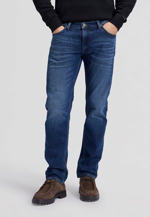 Homme portant un jean bleu, un pull noir et des chaussures en daim marron, les mains dans les poches, debout sur un fond clair uni.