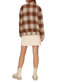 Bruin en crème geruite oversized jas met een textuurafwerking, gedragen over een lichte rok en beige enkellaarsjes. Heeft een kraag en een elastische zoom.