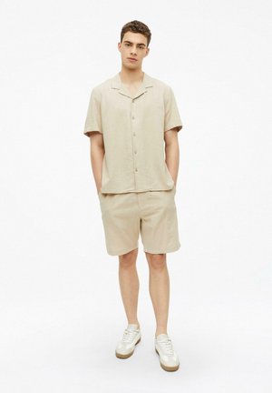 Junger Mann steht mit den Händen in den Taschen, trägt ein beige kurzärmeliges Hemd zum Knöpfen, passende Shorts und weiße Turnschuhe vor weißem Hintergrund.